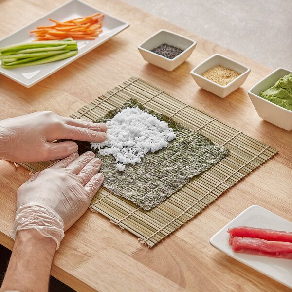 Person making sushi with rice on nori, surrounded by ingredients like vegetables, seeds, and fish on a bamboo mat.