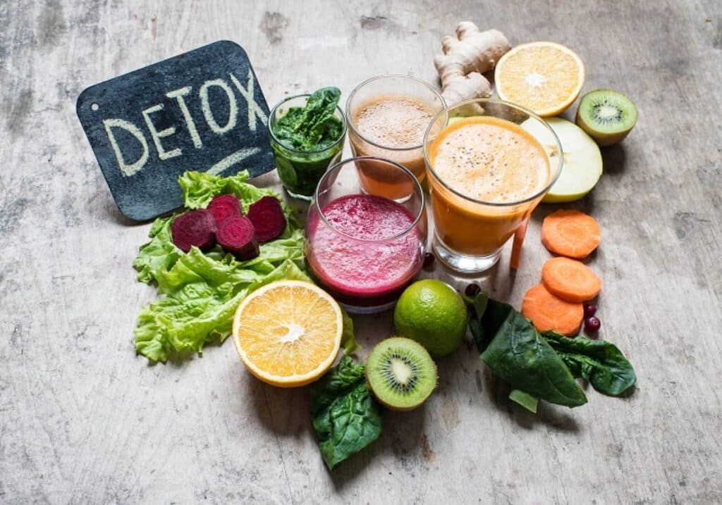 Detox juices with fresh fruits and vegetables on a wooden surface, including oranges, beets, carrots, and greens.