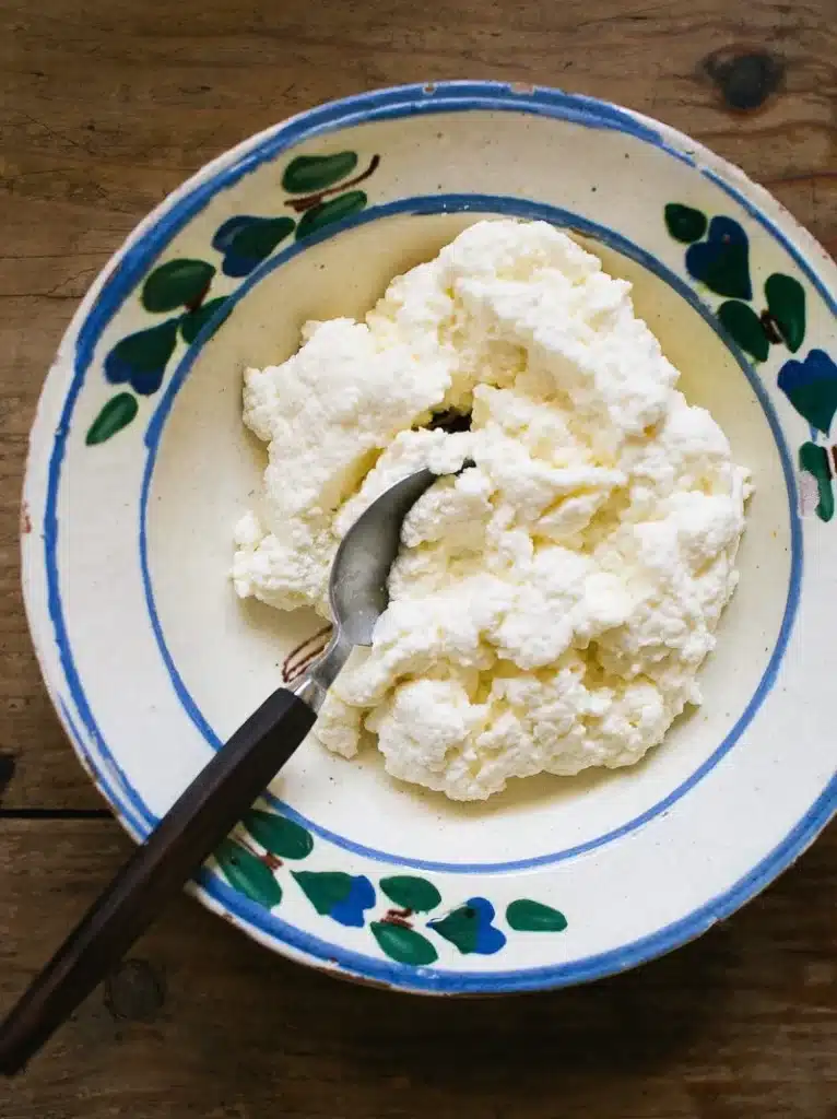Creamy ricotta cheese in a decorative bowl with a spoon on a rustic wooden table.
