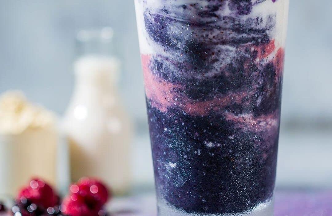 Vibrant purple and pink smoothie in a glass, with milk and berries in the background.