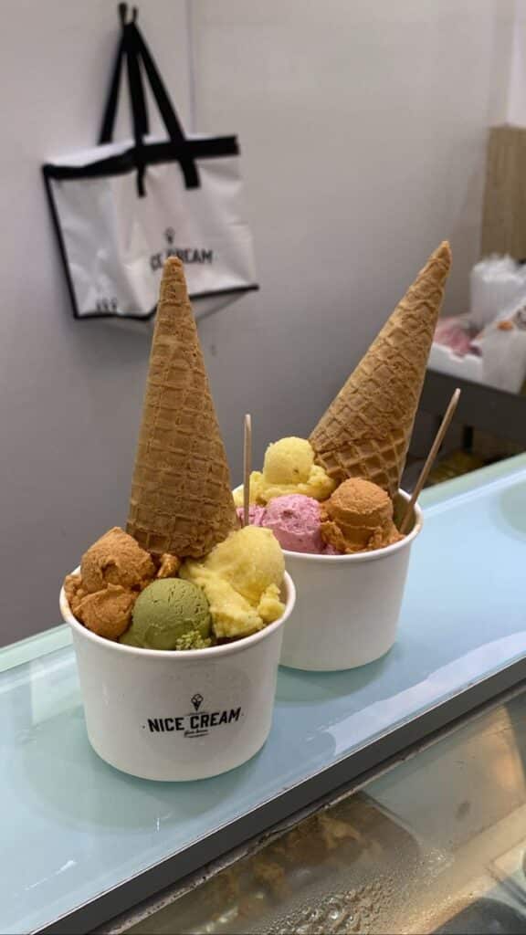Two cups of colorful ice cream scoops with waffle cones, from Nice Cream shop, on a glass counter.