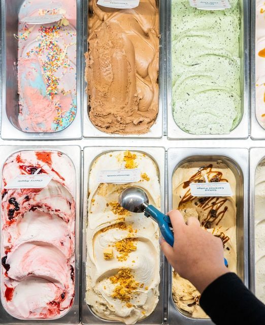 Variety of ice cream flavors in a display case, with a hand scooping one flavor with a blue scoop.