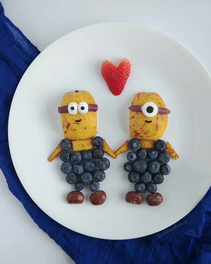 Two fruit characters made from bananas and blueberries holding hands under a strawberry heart on a white plate.