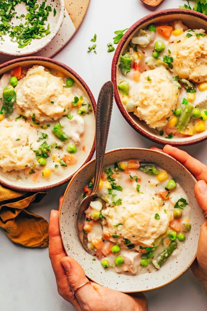 Bowls of creamy vegetable pot pie with flaky biscuits, garnished with fresh herbs, served on a white table.