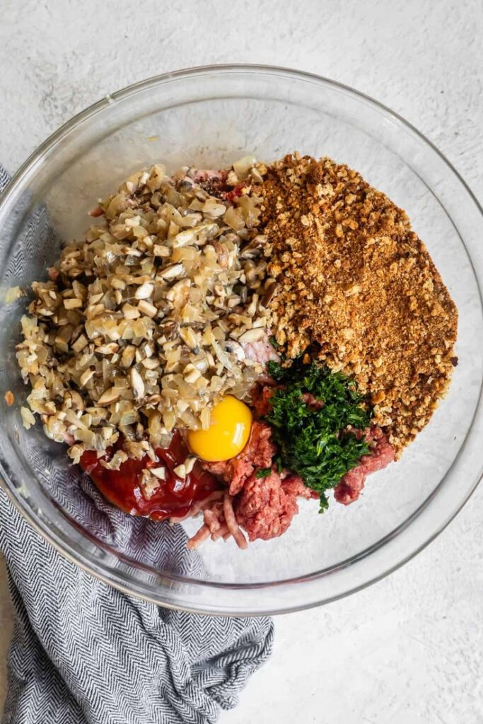 Mixing meatloaf ingredients: ground beef, breadcrumbs, egg, mushrooms, parsley, and ketchup in a bowl.