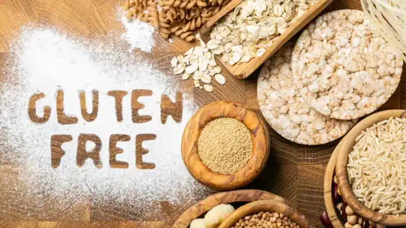 Gluten-free foods arranged on a wooden table with flour spelling 'Gluten Free' surrounded by rice cakes and grains.