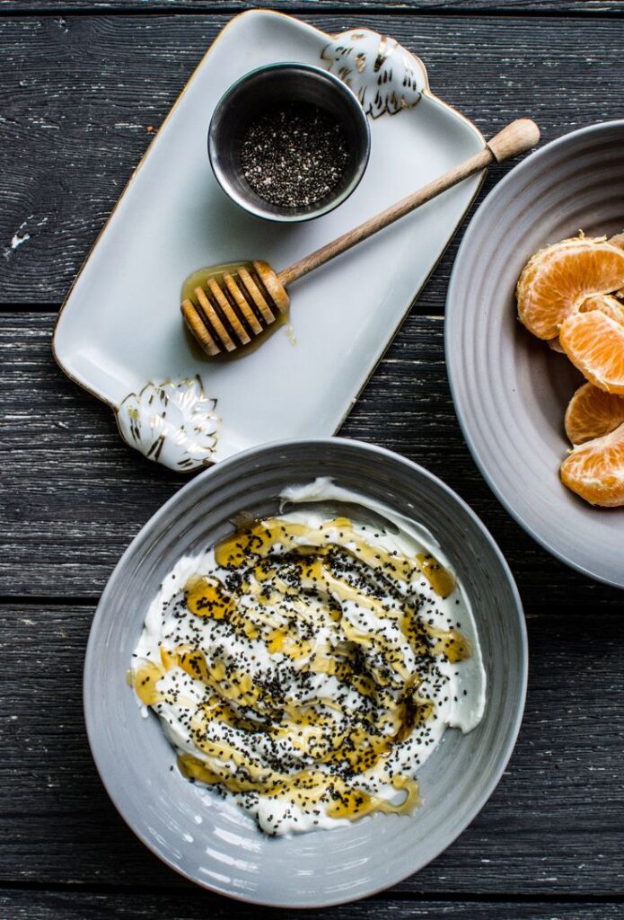 Creamy yogurt with honey and chia seeds in a bowl, alongside sliced oranges on rustic wooden table.