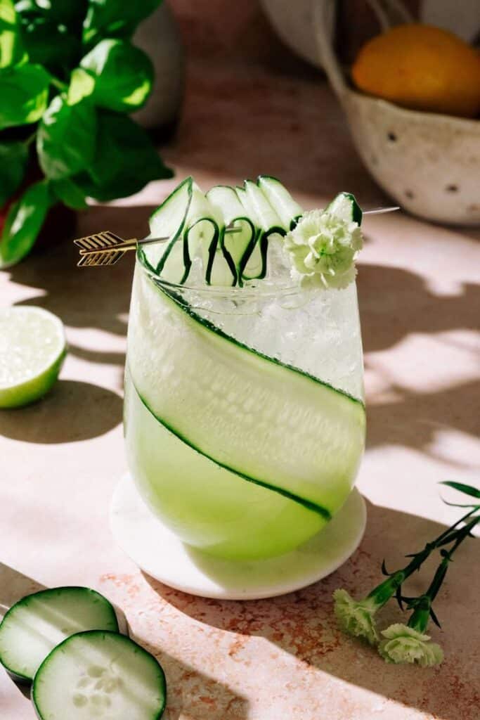 Refreshing cucumber cocktail with sliced garnish and a floral accent, set on a sunlit table with fresh ingredients.