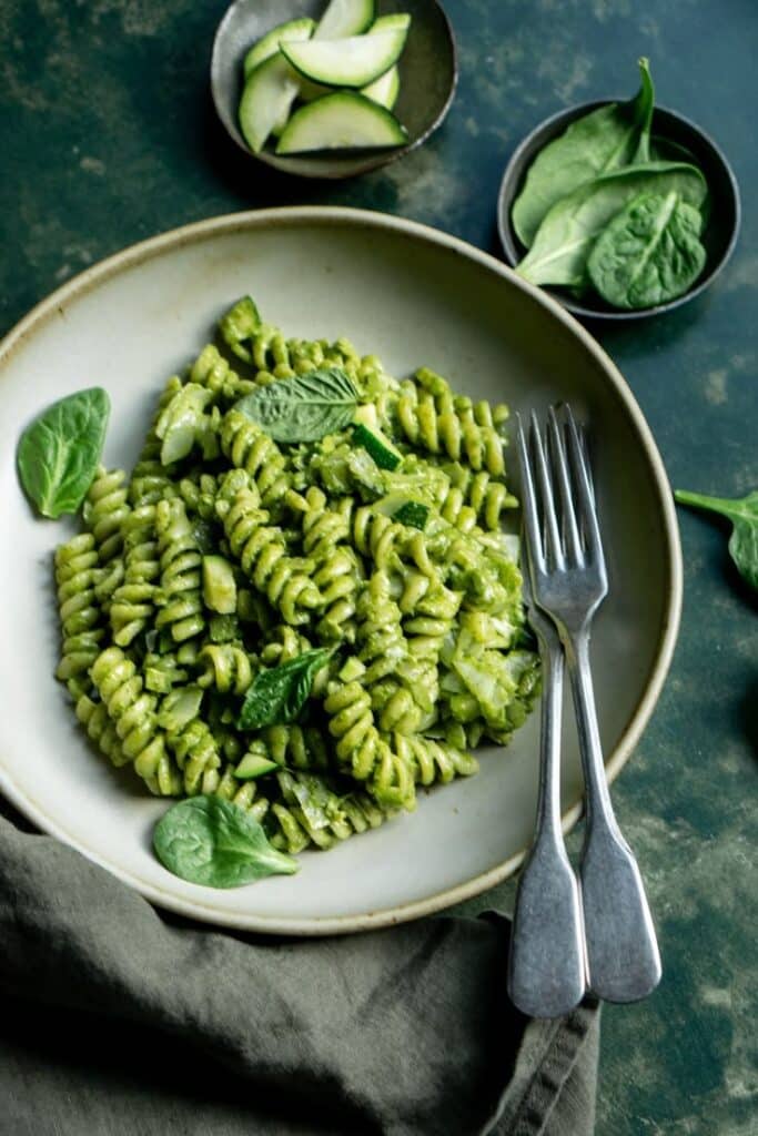 Plate of fusilli pasta with green pesto sauce, garnished with spinach and zucchini slices.