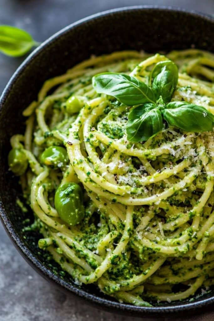 Bowl of spaghetti with fresh pesto, green olives, and basil leaves, topped with grated Parmesan cheese.