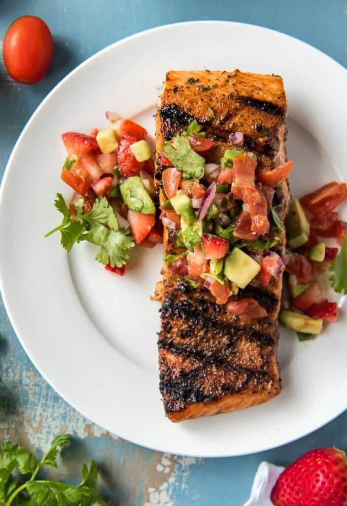 Grilled salmon with fresh avocado salsa on a white plate, garnished with cilantro and tomatoes.