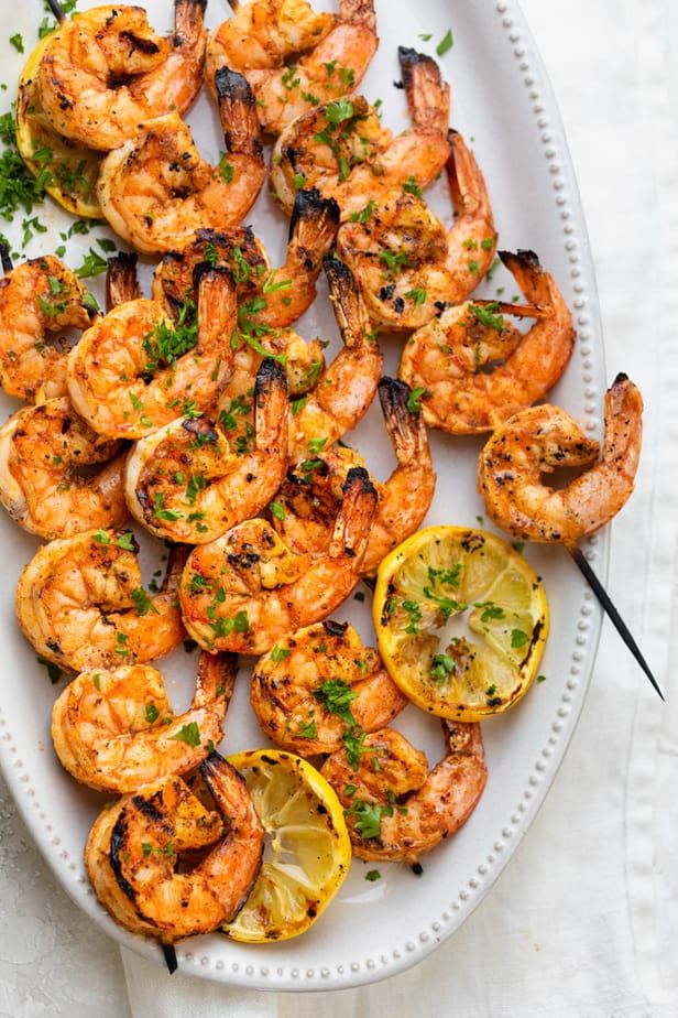 Grilled shrimp skewers with lemon slices and herbs on a white platter.