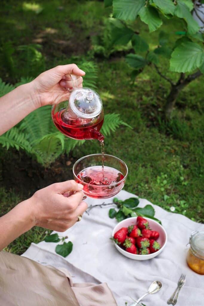 Pouring red herbal tea into glass cup outdoors, strawberries and green leaves on picnic blanket.