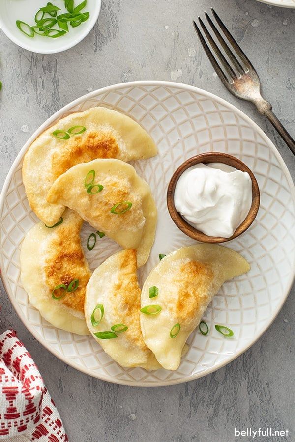 Plate of golden-brown pierogies with sour cream and green onions on a patterned dish, fork on the side.