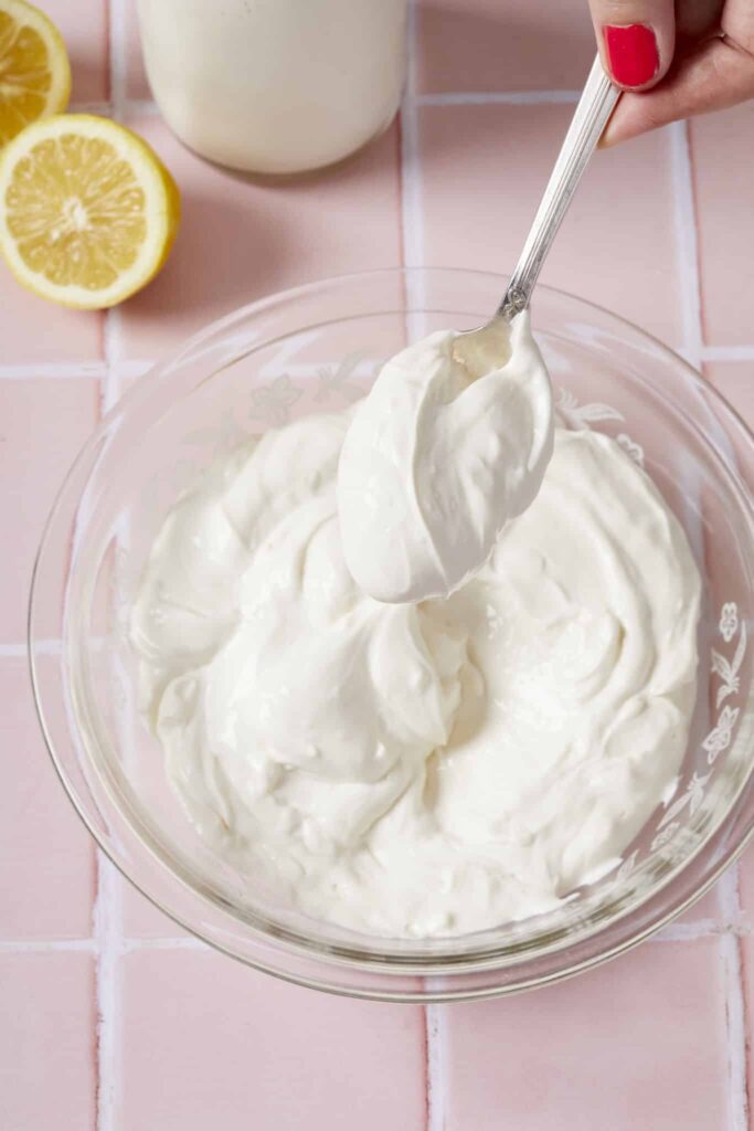 Spoonful of creamy homemade mayonnaise in glass bowl with sliced lemon on pink tile background.