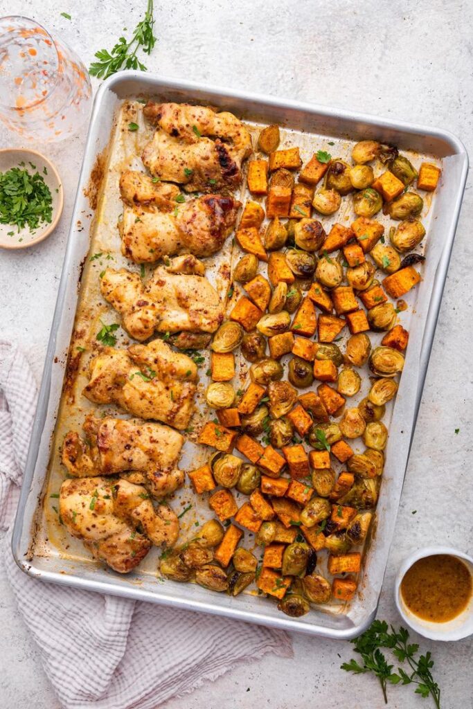 Sheet pan with roasted chicken, sweet potatoes, and Brussels sprouts, garnished with herbs, on a light background.