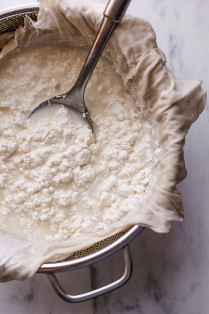 Straining homemade ricotta cheese in a cloth-lined colander with a slotted spoon.