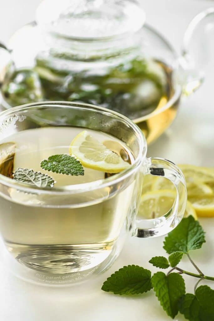 Glass of herbal tea with lemon slices and mint leaves, accompanied by a teapot in the background.