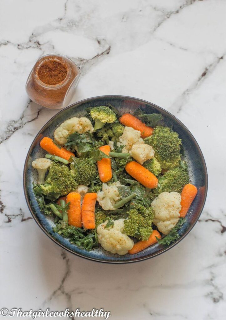 Bowl of fresh steamed vegetables with carrots, broccoli, cauliflower, and a spice jar on a marble surface.