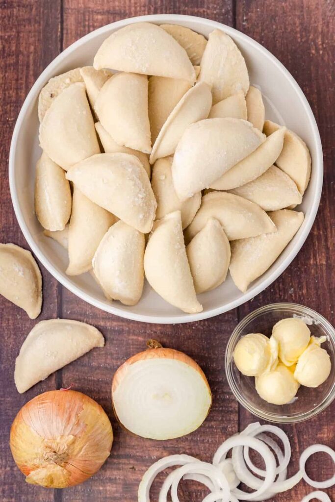 Plate of frozen pierogi with onion slices and butter on wooden table.