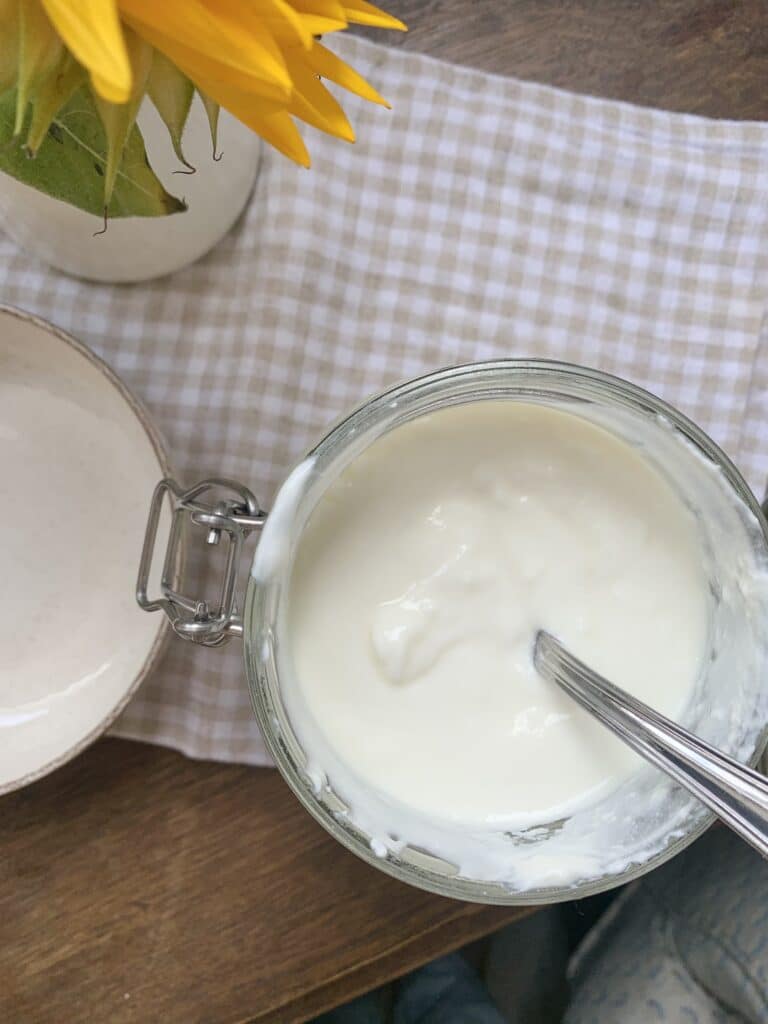 Jar of yogurt with a spoon on a checkered cloth next to a sunflower in a vase.