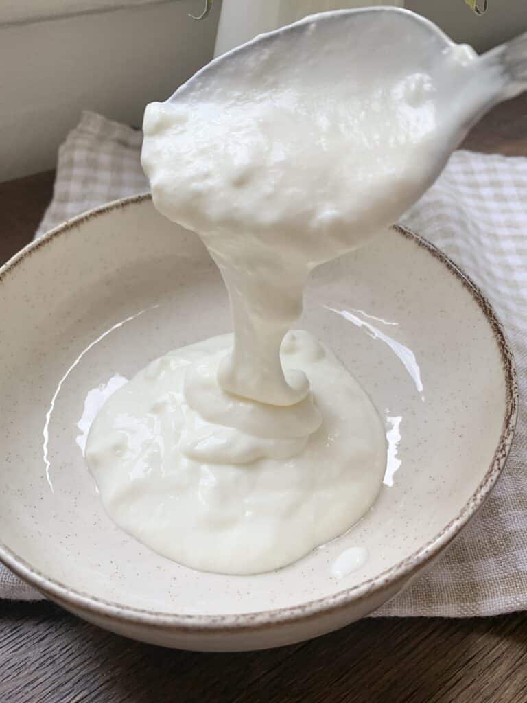 Spoon pouring creamy yogurt into ceramic bowl on a wooden table, natural light, rustic kitchen setting.