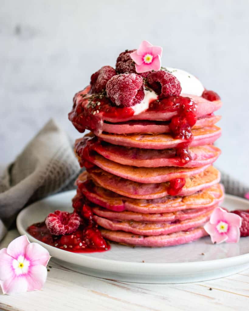 Stack of pink pancakes topped with raspberries and flowers on a white plate.