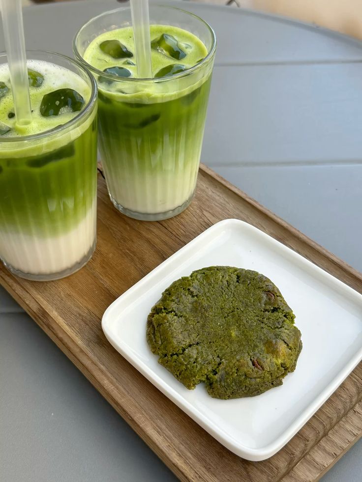 Two iced matcha lattes with a matcha cookie on the side, served on a wooden tray. Refreshing green tea drinks.