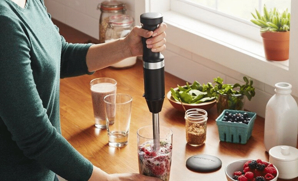 Person blending berry smoothie with hand blender in kitchen. Ingredients: spinach, chia seeds, blueberries, raspberries.