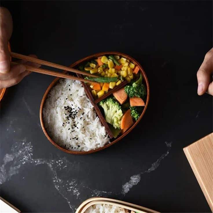 Bento box with rice, sesame seeds, mixed vegetables, and chopsticks on a dark table. Healthy meal concept.