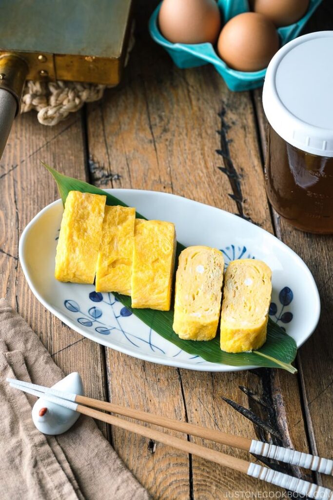 Japanese tamagoyaki slices on a decorative plate with eggs and chopsticks on a wooden table.