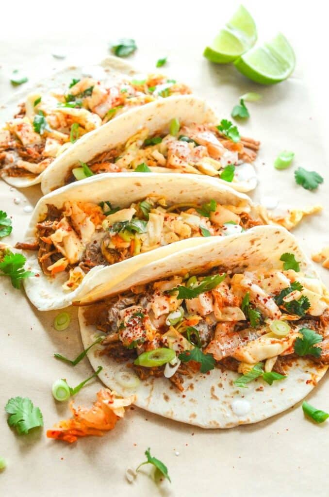 Delicious tacos with fresh cilantro, kimchi, and lime wedges on a parchment background, perfect for a tasty meal.