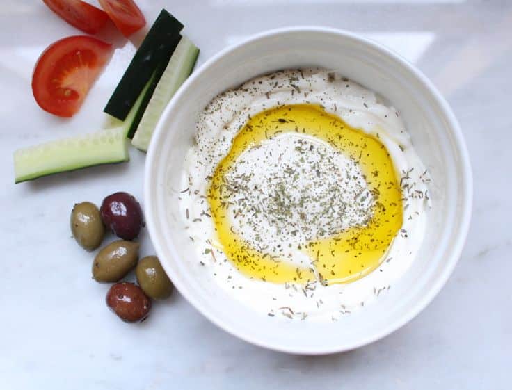 Bowl of tzatziki with olive oil, herbs, olives, cucumber, and tomato slices on a marble surface.