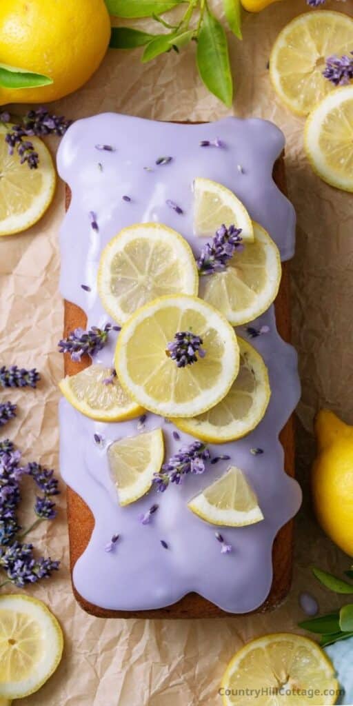 Lavender lemon loaf cake with purple icing, topped with fresh lemon slices and lavender sprigs on parchment.