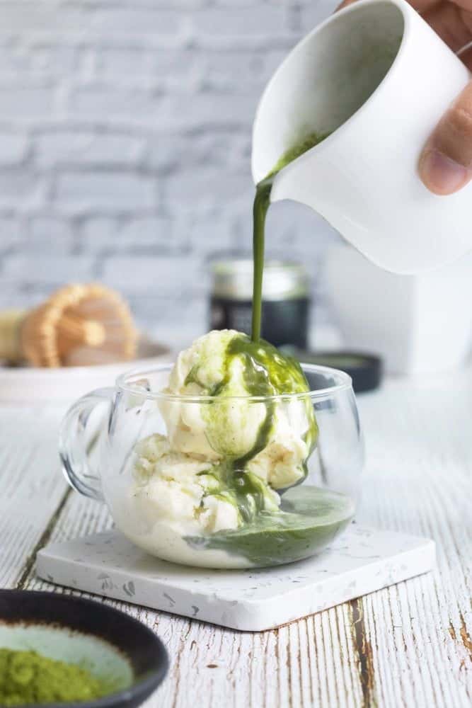 Pouring matcha sauce over vanilla ice cream in a clear glass cup on a wooden table.