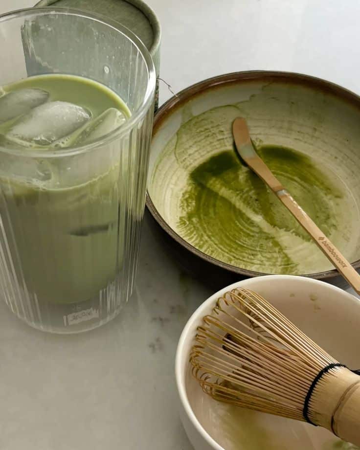 Iced matcha latte with mixing bowl, bamboo whisk, and spoon on a table, perfect for a refreshing drink.