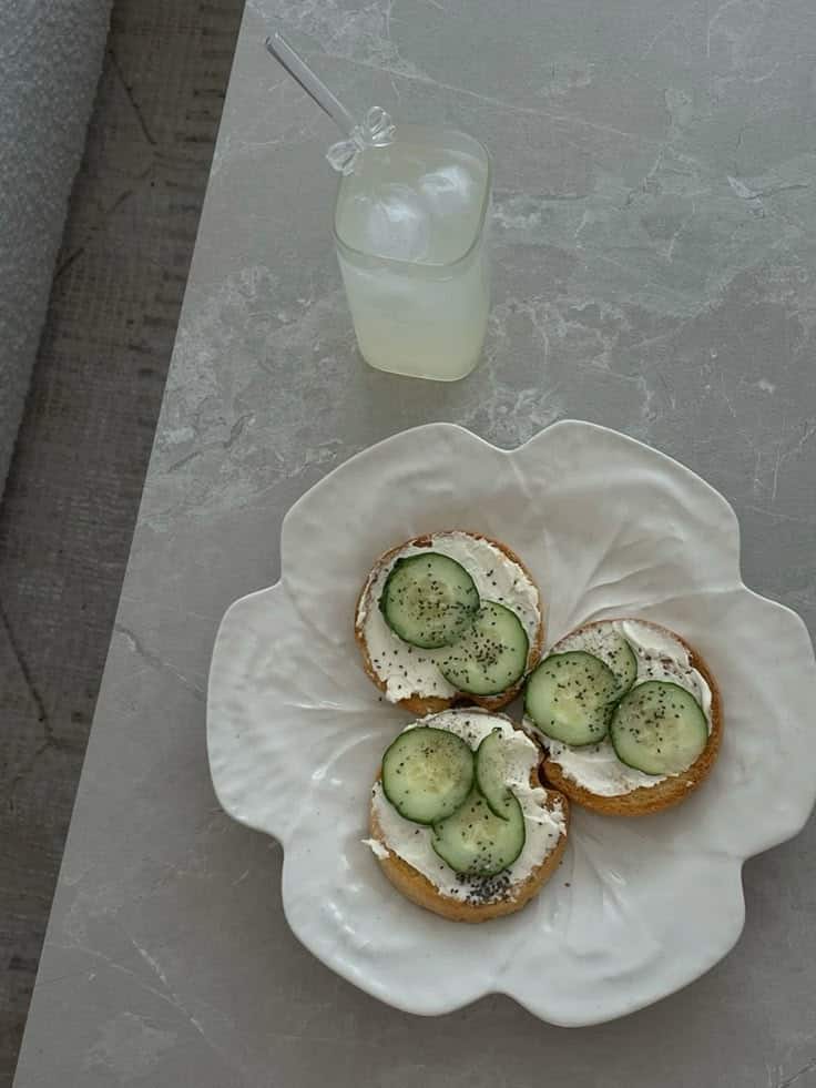 Toasted bagels with cream cheese and cucumber, next to iced lemonade on a marble table.