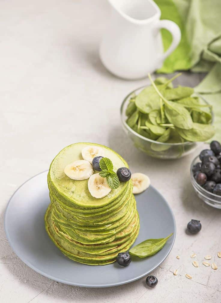 Stack of green spinach pancakes with banana slices and blueberries on a plate, next to fresh spinach and berries.
