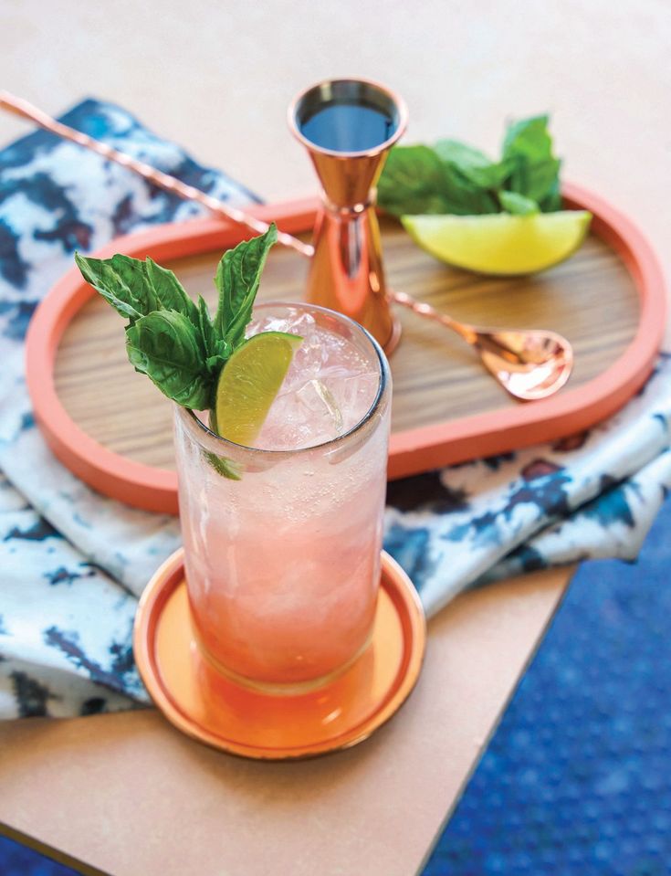 Refreshing pink cocktail with mint and lime garnish, set next to a jigger and serving tray on a patterned cloth.
