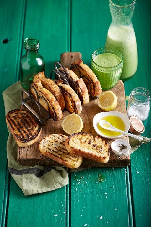 Toasted bread with lemon slices, oil, and a green smoothie on a wooden board against a vibrant green background.