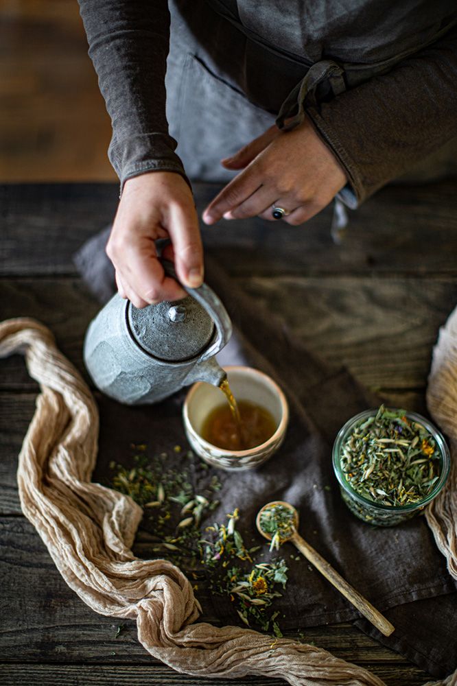 Person pouring herbal tea from a teapot into a cup on a wooden table with loose herbs and a spoon nearby.