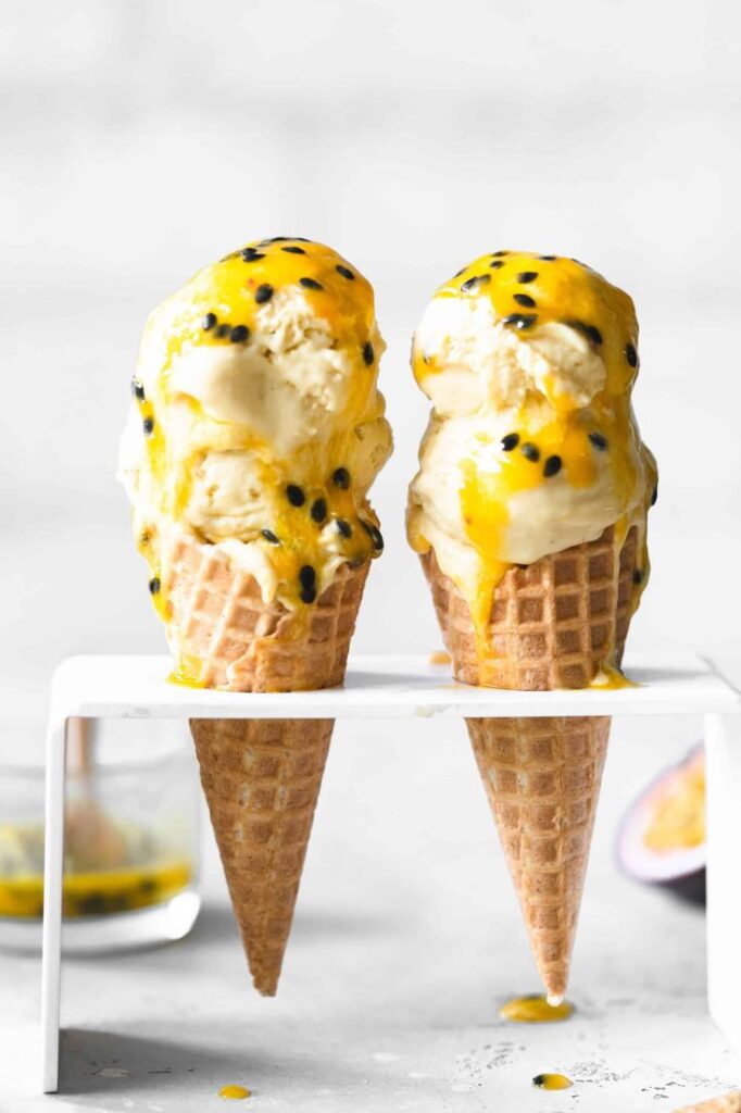 Two waffle cones with passion fruit ice cream topped with vibrant yellow sauce and seeds, on a white stand.