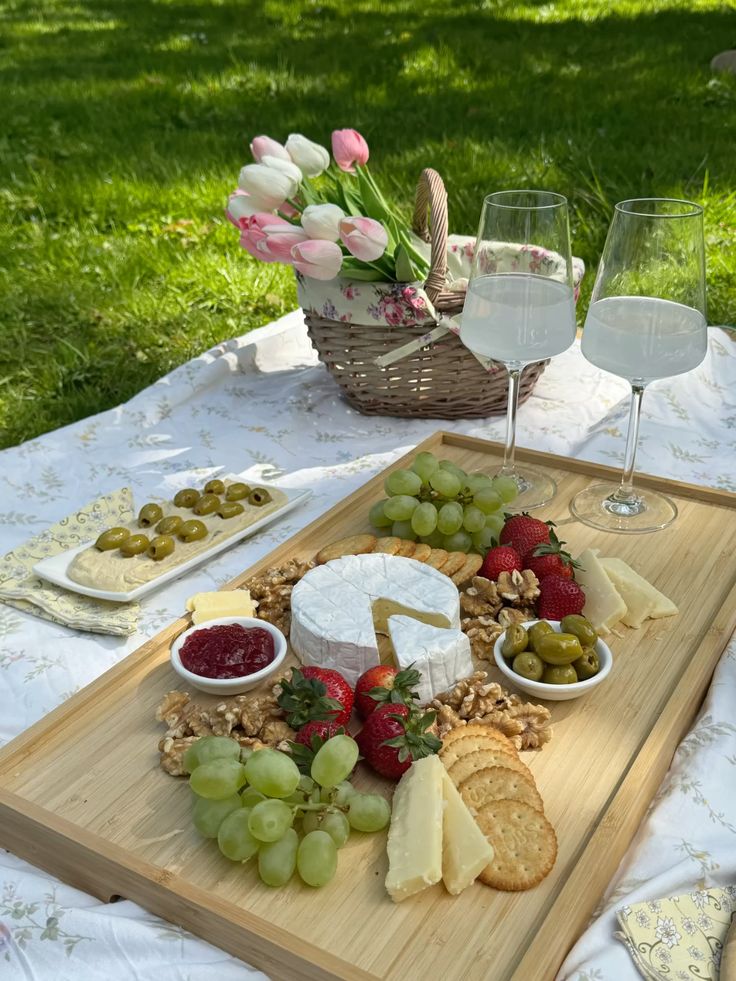 Picnic setup with cheese board, fruits, crackers, wine glasses, and tulips on a blanket in a sunny park.