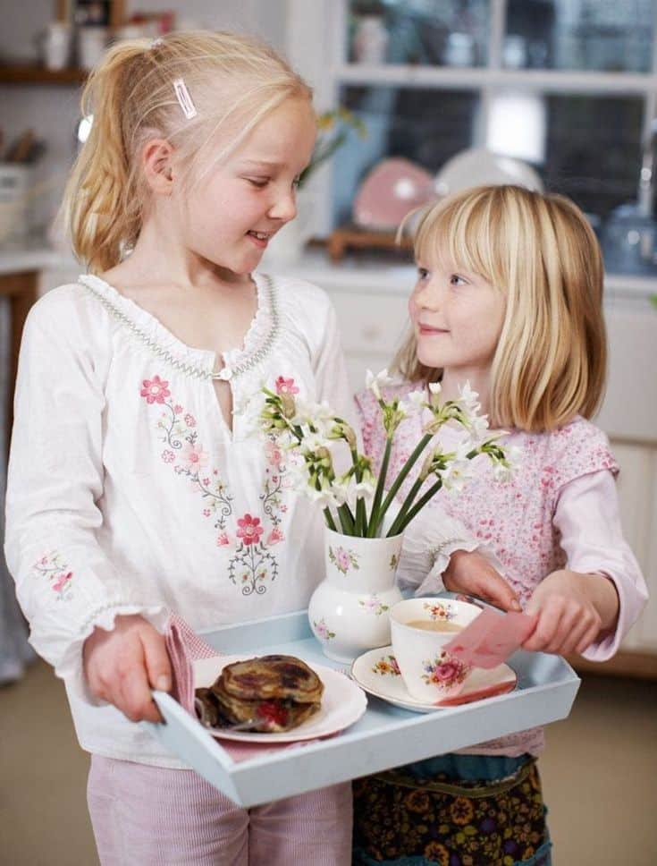 Two children with a breakfast tray, including pancakes, tea, and flowers, sharing a moment in a cozy kitchen setting.