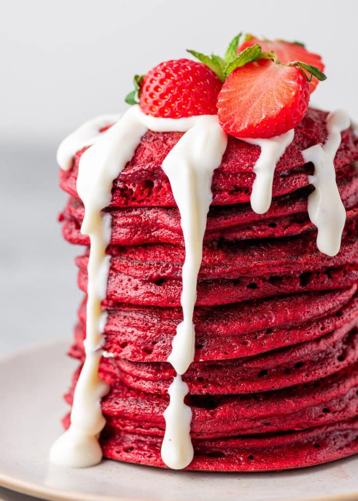 Stack of red velvet pancakes with cream cheese frosting and strawberries on top.