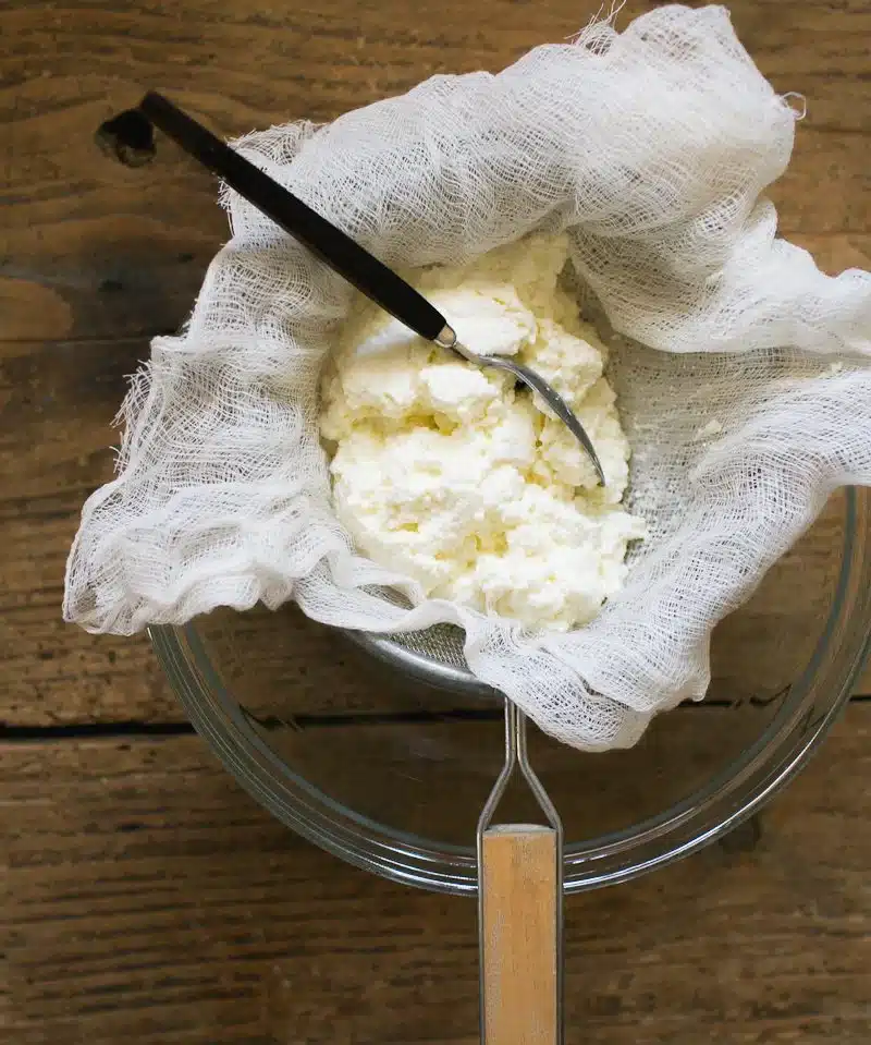 Homemade ricotta cheese in a cheesecloth with spoon on wooden background, fresh dairy product preparation.