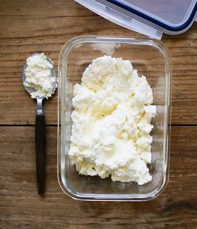 Homemade whipped ricotta in a glass container on a wooden table with a spoonful beside it.