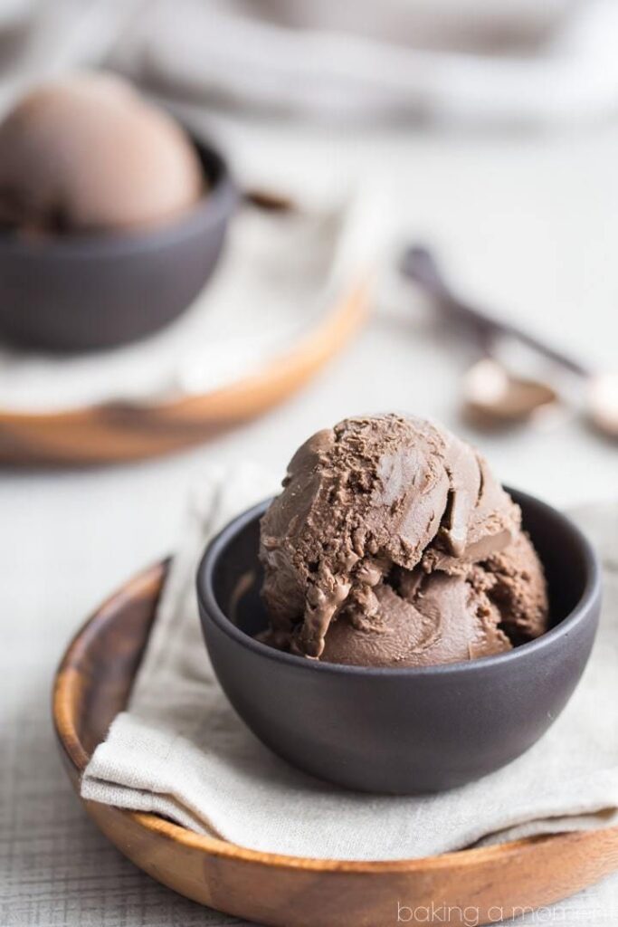 Rich chocolate ice cream in a dark bowl on a wooden plate, with a blurred second serving in the background.