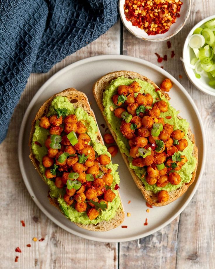 Avocado toast topped with spicy roasted chickpeas and herbs on a white plate, garnished with sliced green onions.