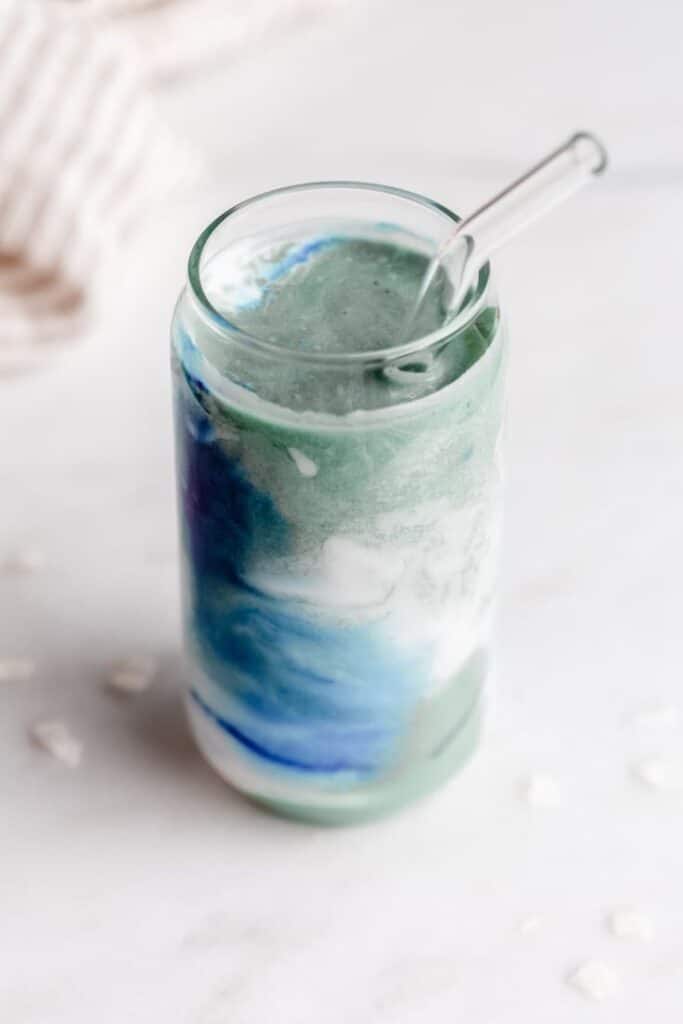 Vibrant blue and green smoothie in a clear glass with straw, placed on a light marble surface.